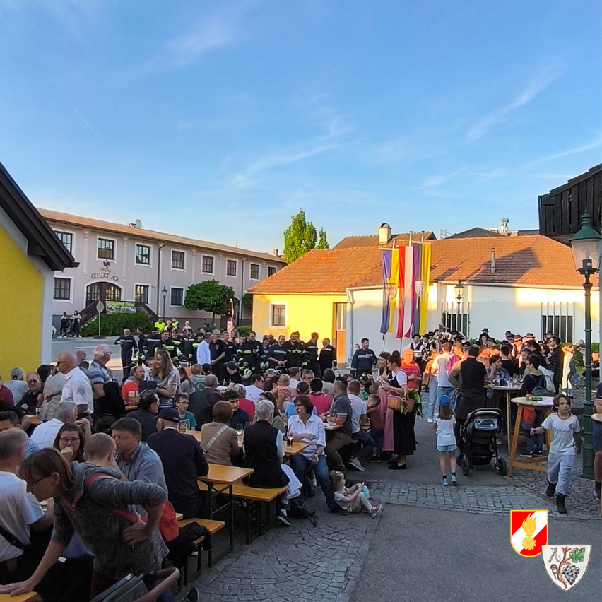 Eine große Menschenmenge versammelt sich um Tische in einem Außenbereich. Es gibt Gebäude mit Fahnen, und der Himmel ist blau mit einigen Wolken. Ein Kind geht auf der Kopfsteinpflasterstraße.