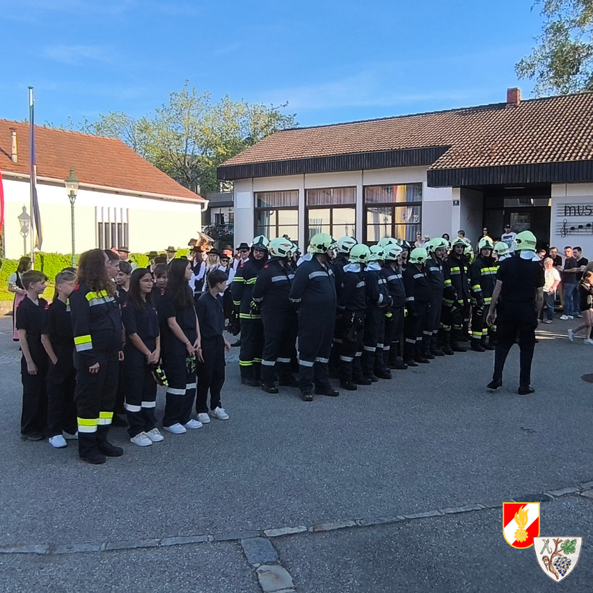Eine Gruppe von Feuerwehrleuten in voller Uniform, einschließlich Kindern, steht in einer Reihe vor der Feuerwache, wobei eine Flagge und das Wappen des Gebäudes im Hintergrund zu sehen sind.