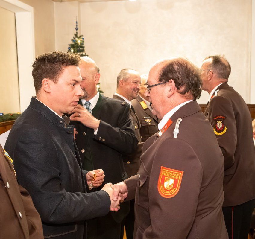 Zwei Männer in Uniformen schütteln sich die Hände in einem Raum mit anderen. Ein Weihnachtsbaum steht hinter ihnen.
