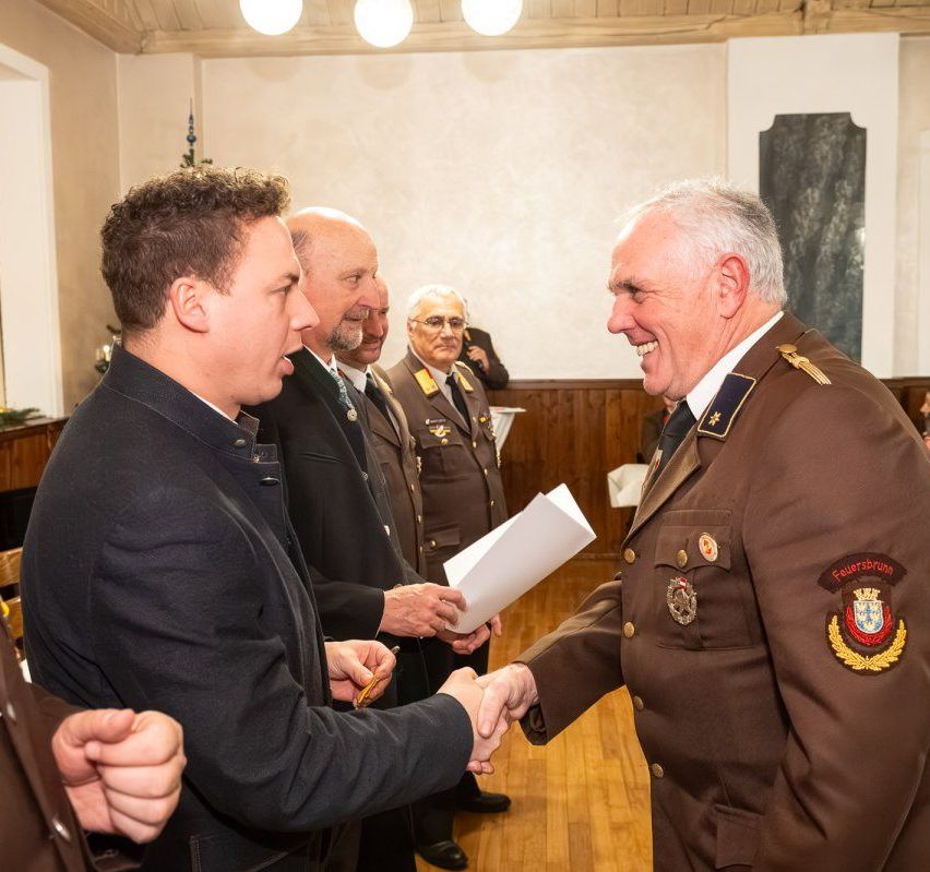 Ein Militäroffizier in Uniform überreicht einem Mann im Anzug ein Papier, während andere zusehen. Sie sind drinnen, mit Holzwänden und einem Schreibtisch im Hintergrund.