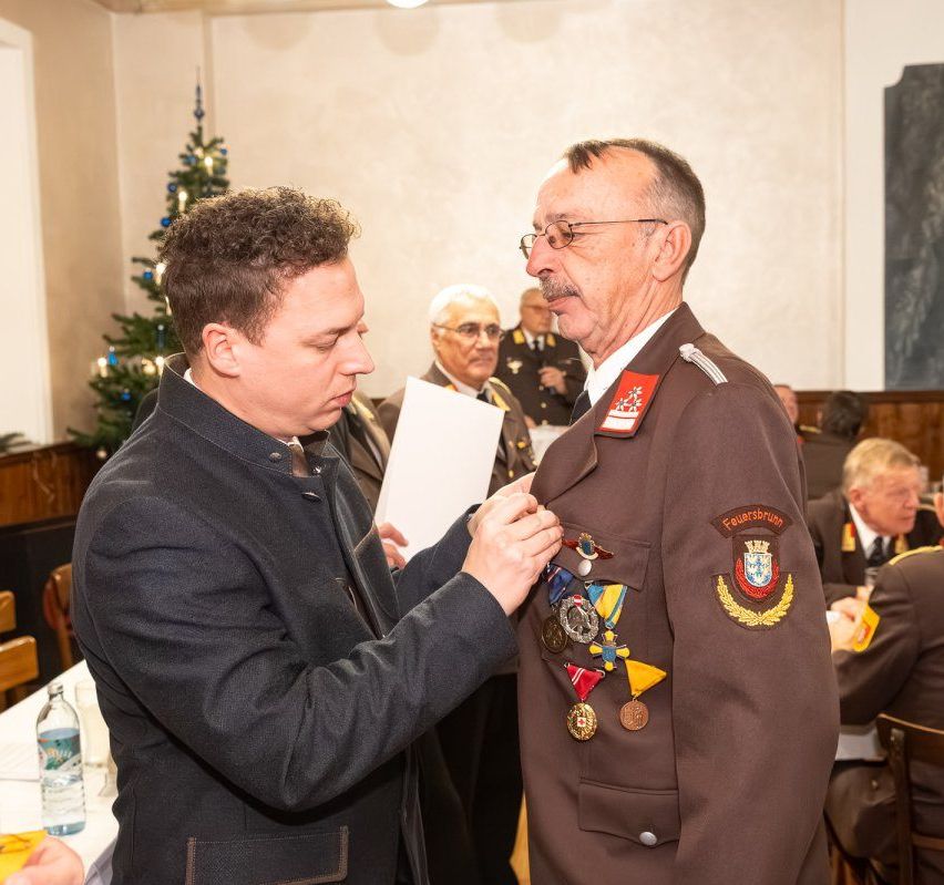 Ein Mann befestigt Medaillen an der Uniform eines anderen Mannes. Der Mann mit Medaillen trägt eine braune Uniform mit einem Abzeichen auf der linken Schulter. Der Raum ist mit einem Weihnachtsbaum dekoriert und einige Leute sitzen auf Stühlen.