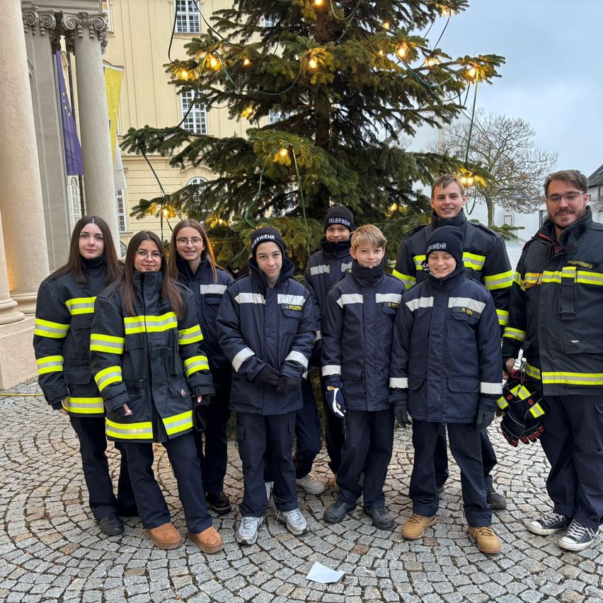 Eine Gruppe von Feuerwehrleuten posiert für ein Foto vor einem geschmückten Weihnachtsbaum, mit einem historischen Gebäude und Bäumen im Hintergrund.