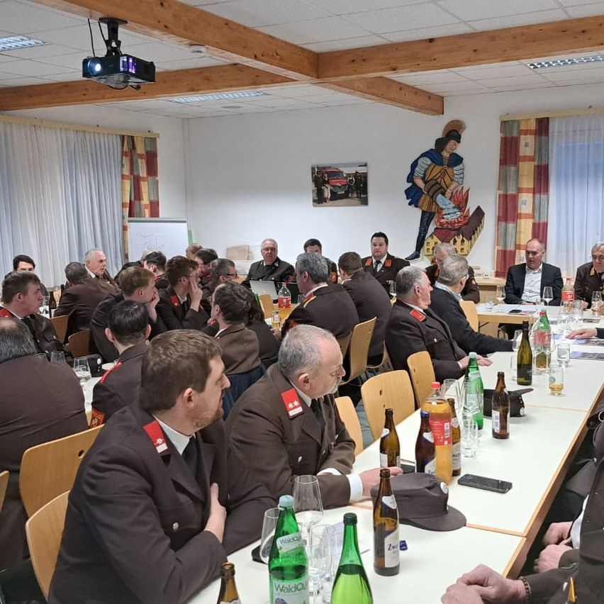 Eine Gruppe von Menschen in formeller Kleidung sitzt um einen Tisch mit Flaschen und Gläsern herum. Eine Leinwand und eine Tafel sind an der Wand. Ein großes Gemälde eines Ritters ist an der Wand.