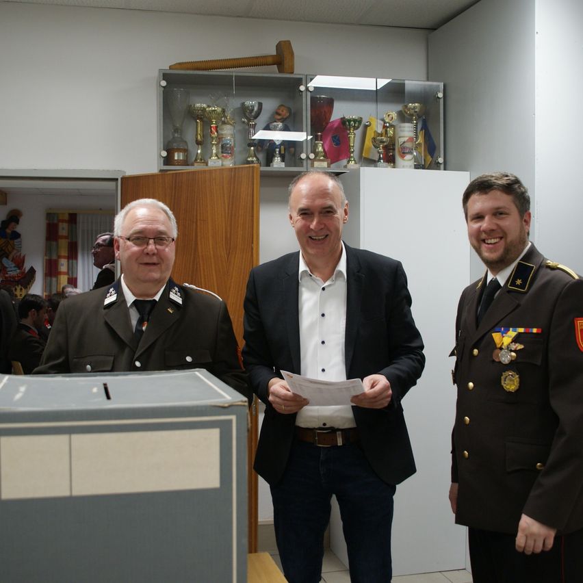 Drei Männer in Uniformen und Anzügen stehen vor einer Wahlurne. Dahinter ist ein Trophäenschrank mit verschiedenen Trophäen zu sehen.