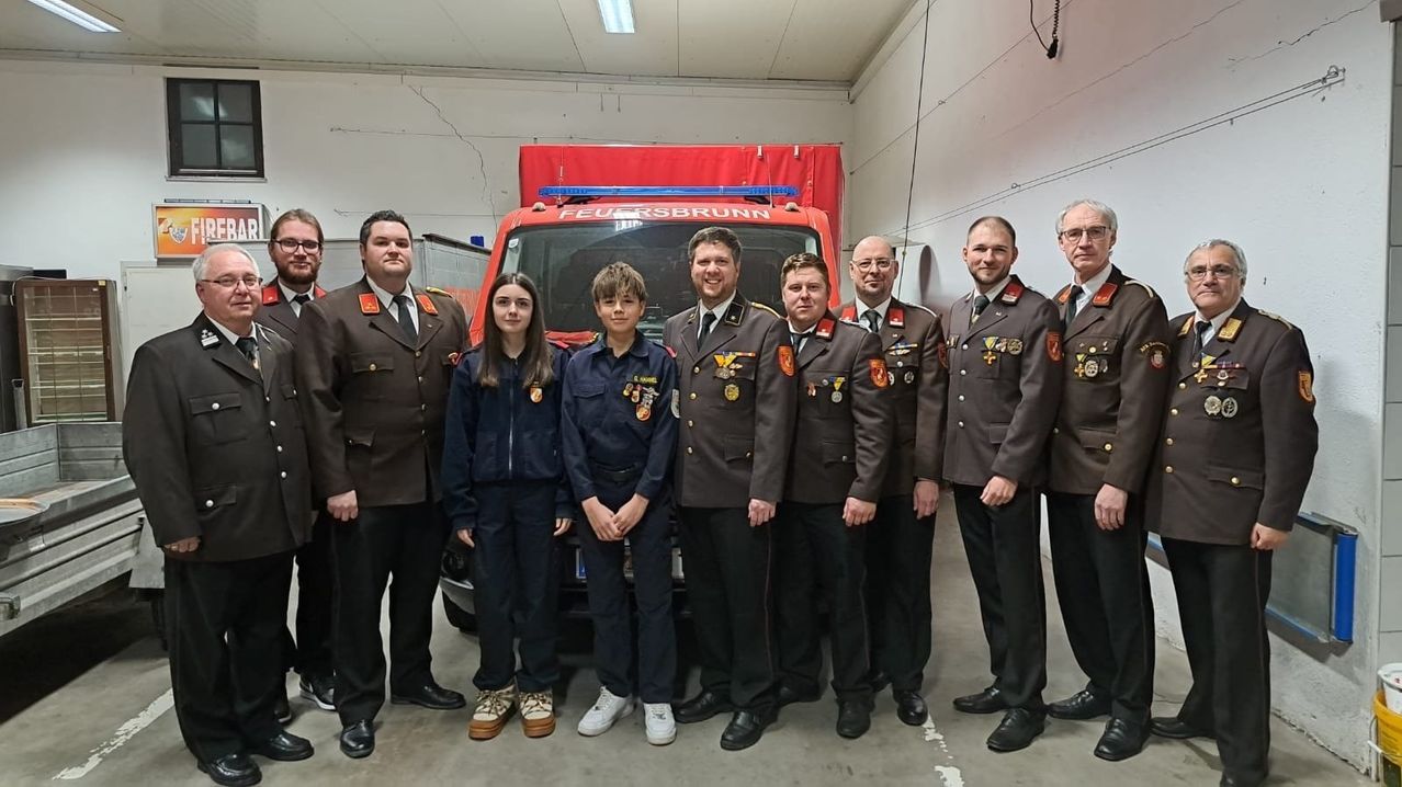 Eine Gruppe von Feuerwehrleuten und zwei Kindern steht vor einem roten Feuerwehrauto in einer Garage. Sie tragen Uniformen.