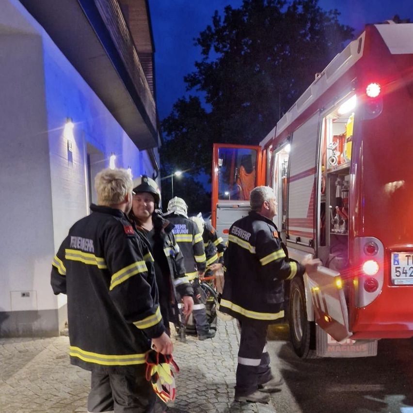 Firefighters are standing by a red fire truck at night, with one man talking to another and another holding a helmet.