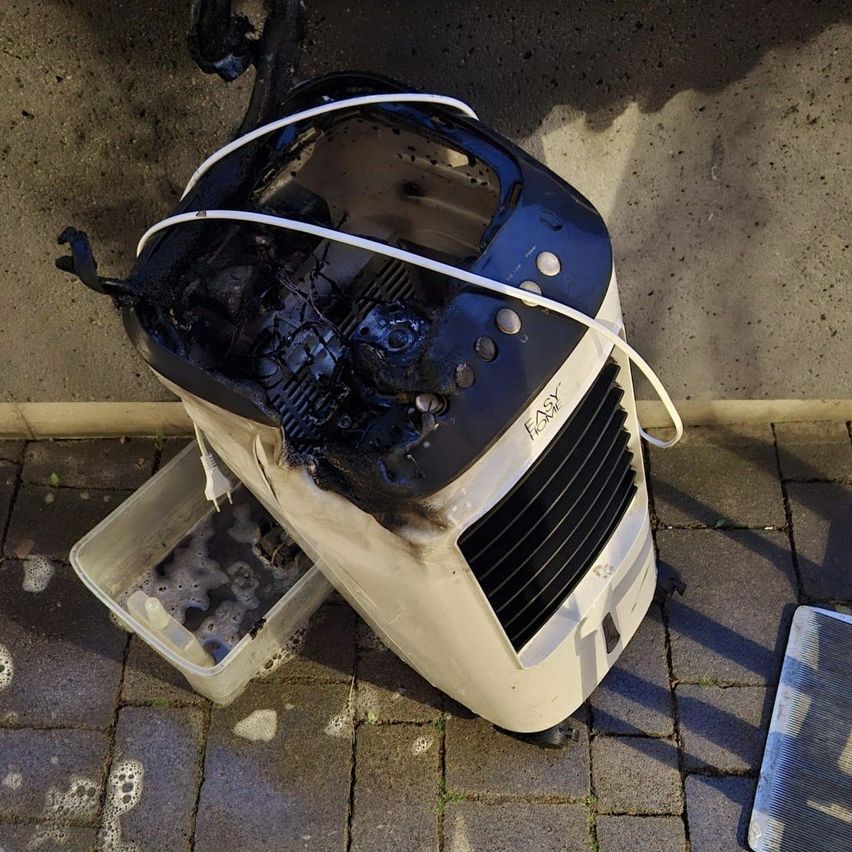 A broken air cooler lies on the ground with a damaged casing, burnt wires, and an open container of water nearby.