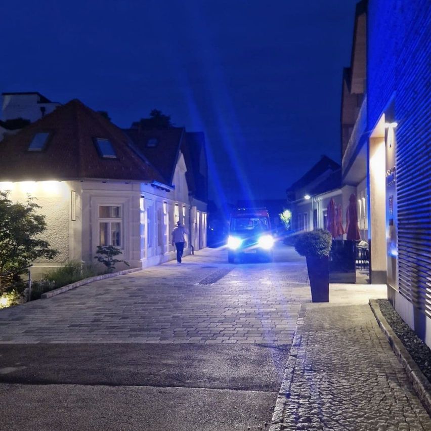 A nighttime scene shows a person walking along a street with illuminated buildings and a parked vehicle with its headlights on.