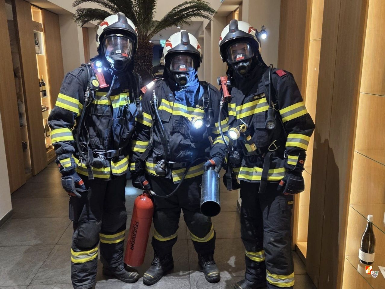 Three firefighters stand together wearing full gear, helmets, and masks. One holds a fire extinguisher, another holds a large pipe, and the third wears a blue scarf. They are in a well-lit corridor.