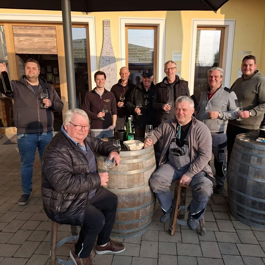 A group of men are gathered around a barrel, posing for a photo with wine glasses and bottles in hand. They are smiling and seem to be enjoying themselves.