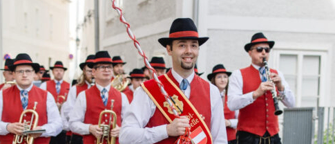 Eine Blaskapelle in traditioneller österreichischer Kleidung tritt auf der Straße auf, wobei ein Mann eine Flagge hält und andere Trompeten und Klarinetten spielen.