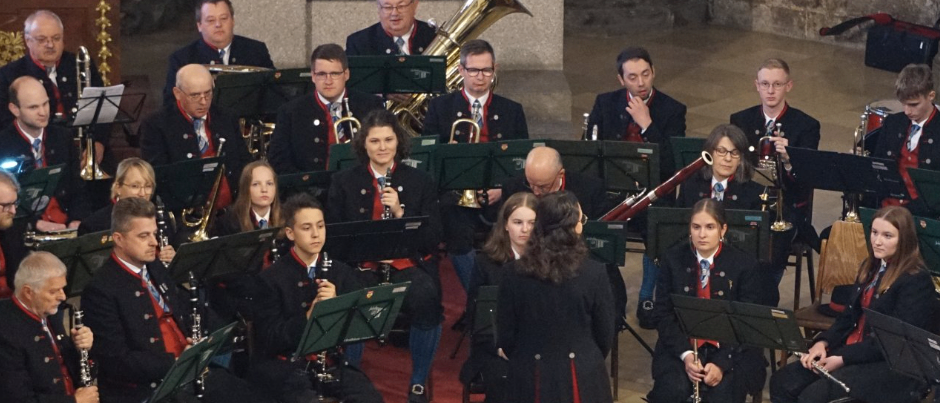 Ein Orchester mit verschiedenen Instrumenten, einschließlich Tuba und Posaune, spielt in einem Raum mit Holzboden. Einige tragen Brillen und Krawatten.