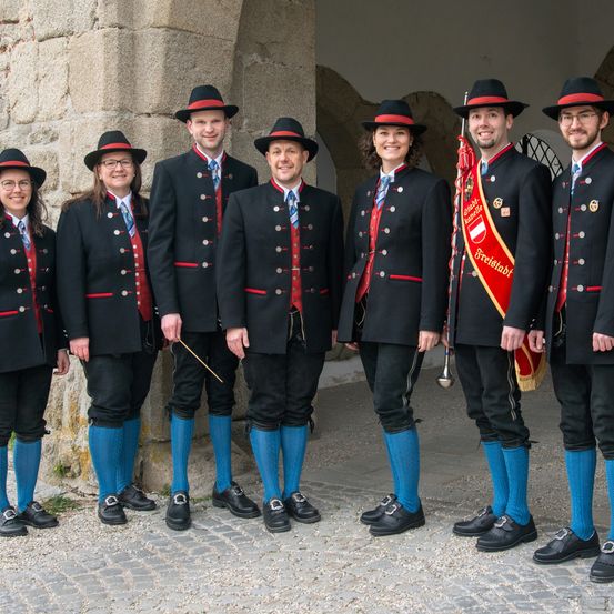 Eine Gruppe von formell gekleideten Menschen, möglicherweise ein Marschkapelle, steht vor einem Steinbogen, alle tragen schwarze Uniformen mit roten Details und blauen Socken.