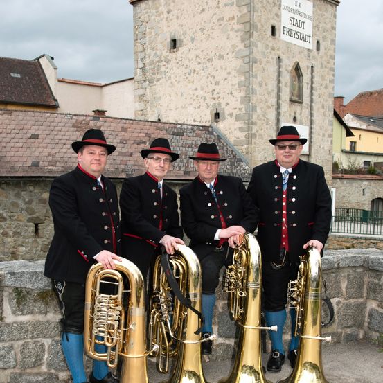 Vier Männer in traditionellen Uniformen stehen vor einem historischen Gebäude mit einem Schild, das 'Stadt Freistadt' besagt. Sie halten große Messinginstrumente und scheinen Teil einer Blechbläserkapelle zu sein.