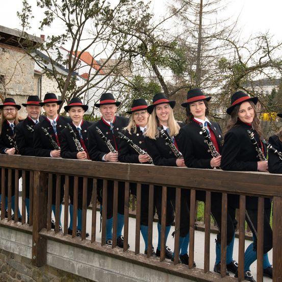 Eine Gruppe von Menschen in traditioneller Kleidung posiert mit Klarinetten auf einer Brücke. Der Hintergrund zeigt Bäume und Gebäude.