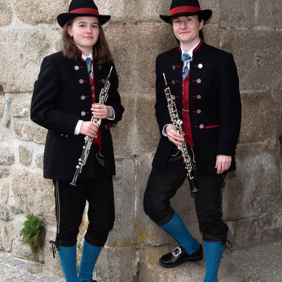 Zwei Personen in traditioneller Kleidung, jede mit einer Klarinette, stehen an einer Steinmauer, möglicherweise bei einer musikalischen Aufführung.