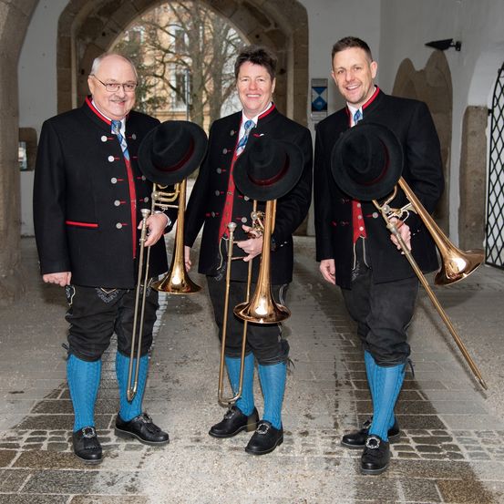 Drei Männer in traditioneller Kleidung posieren mit Posaunen und lächeln. Sie stehen eng beieinander und halten ihre Instrumente mit Hüten auf dem Kopf. Der Hintergrund zeigt ein Portal mit Steinmauern.