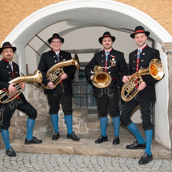 Vier Männer in traditioneller Kleidung, einschließlich Hüten und blauen Socken, stehen zusammen und halten Blechblasinstrumente. Sie stehen unter einem Bogen vor einem Gebäude.