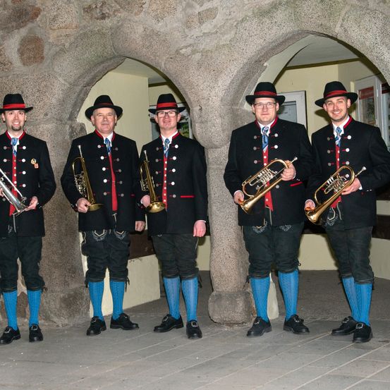 Fünf Männer in traditioneller Kleidung, einschließlich Hüten, Uniformen und blauen Socken, stehen zusammen in einem Bogen und halten Trompeten.