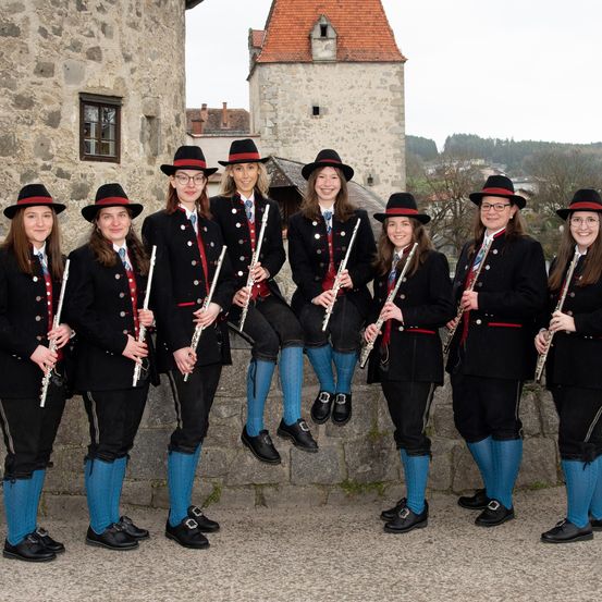 Eine Gruppe von Frauen in schwarzen Uniformen und Hüten posiert für ein Foto mit Flöten vor einer Steinmauer.