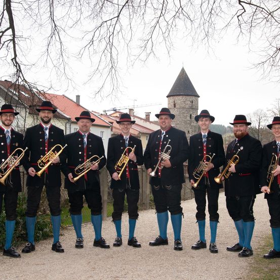 Eine Gruppe von Männern in traditioneller Kleidung posiert mit Trompeten vor einem mittelalterlichen Turm.