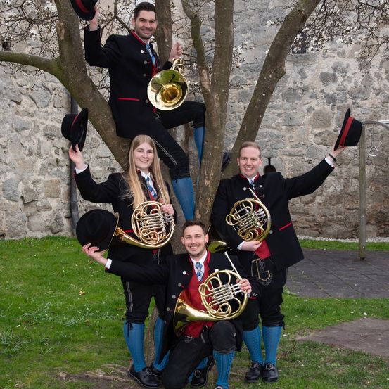 Fünf Musiker posieren mit ihren Posaunen und Hüten in einem Baum, vor einem Steinmauer im Hintergrund.