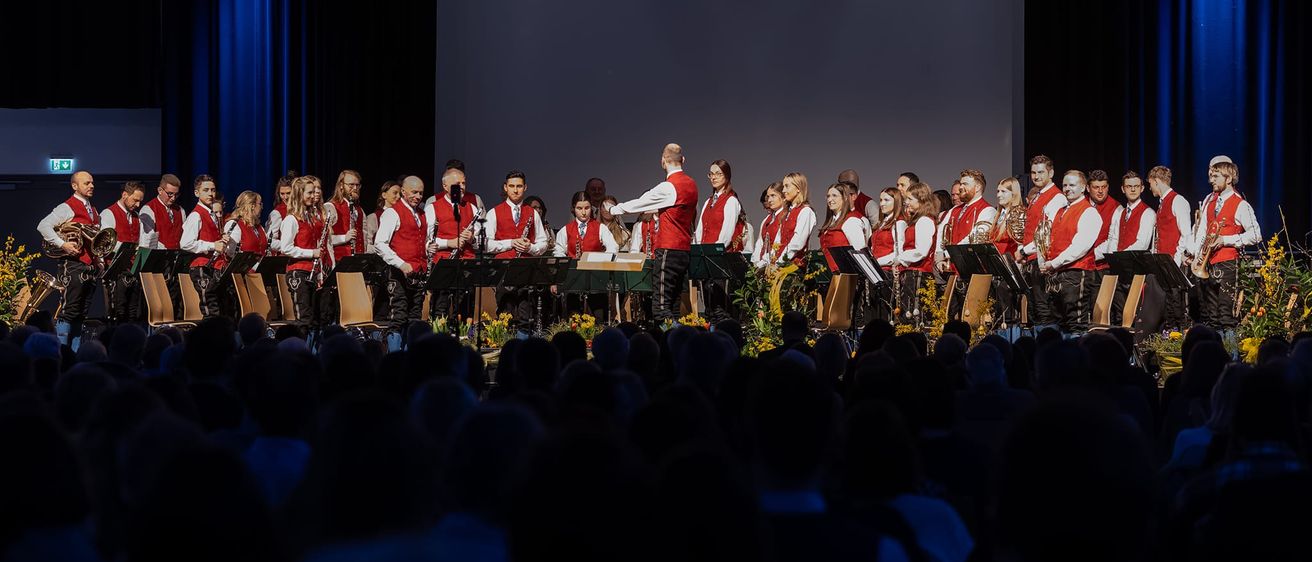 Ein Chor auf der Bühne mit Instrumenten, der vor einem Publikum auftritt. Sie tragen rote Westen und schwarze Hosen.