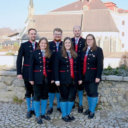 Eine Gruppe von Menschen in traditioneller Kleidung steht vor einem historischen Gebäude. Sie lächeln und scheinen für ein Foto zu posieren.