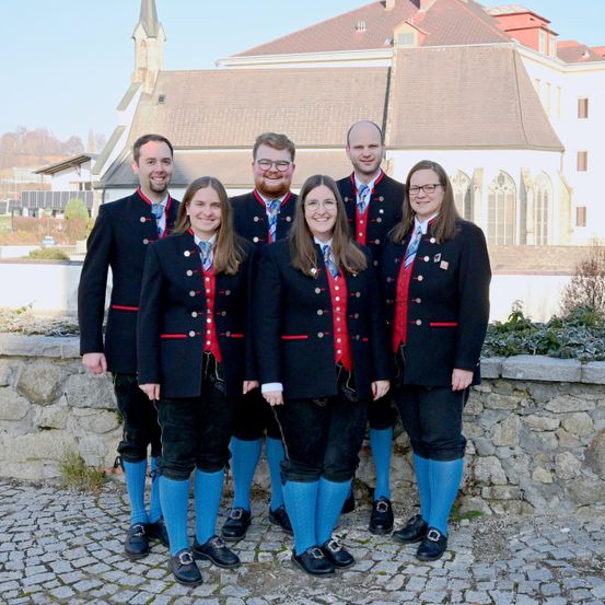 Eine Gruppe von Menschen in traditioneller Kleidung steht vor einem historischen Gebäude. Sie lächeln und scheinen für ein Foto zu posieren.