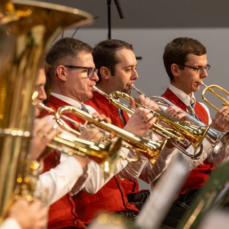 Eine Gruppe von Musikern in roten Westen spielt Trompeten während einer Aufführung. Sie alle schauen in die gleiche Richtung. Der Mann in der Mitte hält einen Posaune.