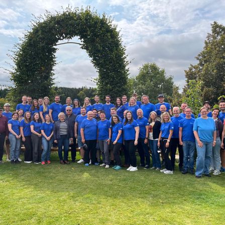 Eine große Gruppe von Menschen in blauen T-Shirts posiert für ein Foto im Park. Sie stehen vor einem grünen Bogen und einem bewölkten Himmel.