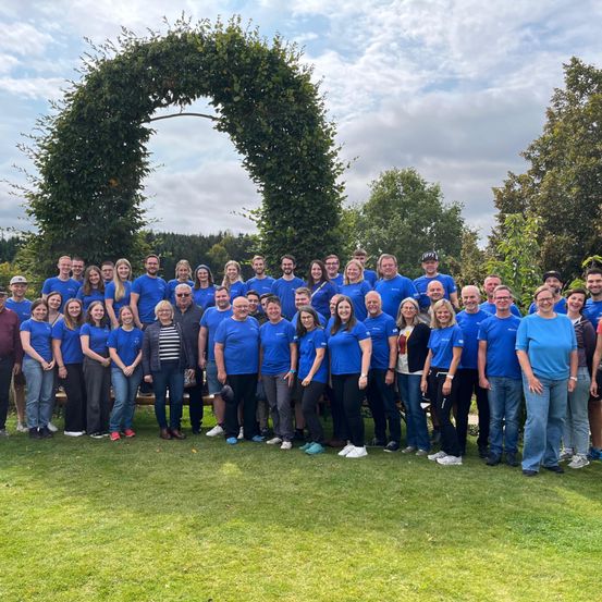 Eine große Gruppe von Menschen in blauen T-Shirts posiert für ein Foto im Park. Sie stehen vor einem grünen Bogen und einem bewölkten Himmel.