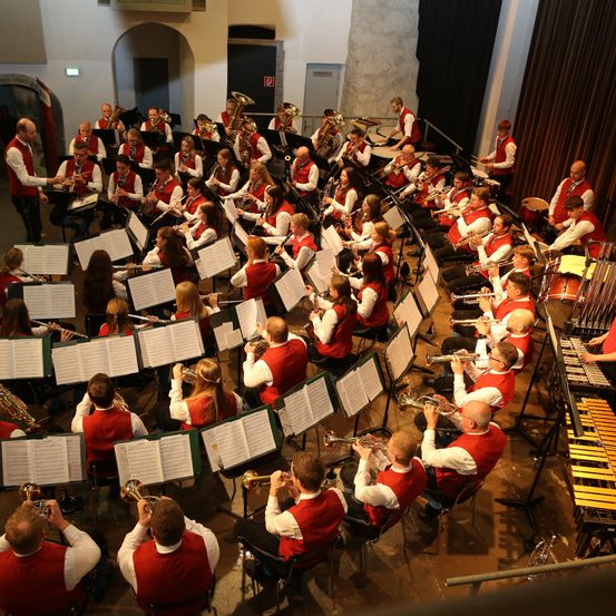 Eine Blaskapelle spielt auf der Bühne mit verschiedenen Instrumenten. Sie tragen passende rote Westen und sitzen in Stühlen. Vor ihnen liegen Musikblätter.