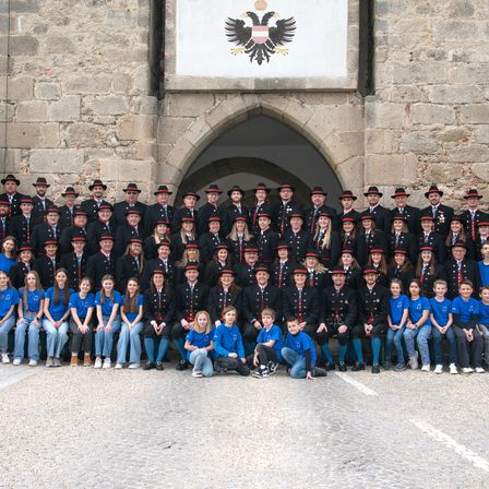 Eine große Gruppe von Menschen, einige in Uniformen, posiert für ein Foto vor einem Torbogen mit einem Adleremblem. Sie sitzen und stehen auf Kopfsteinpflasterboden.