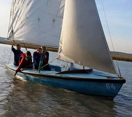 Eine Gruppe von Menschen segelt mit einem blauen Segelboot mit weißer Segel auf einem ruhigen Gewässer. Das Boot hat die Nummer 614 auf seiner Seite.
