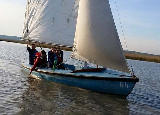 Eine Gruppe von Menschen segelt mit einem blauen Segelboot mit weißer Segel auf einem ruhigen Gewässer. Das Boot hat die Nummer 614 auf seiner Seite.