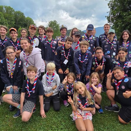 Eine Gruppe junger Pfadfinder, sowohl Jungen als auch Mädchen, posiert für ein Foto auf einer Wiese. Sie tragen ihre Pfadfinderuniformen und einige tragen eine Brille.