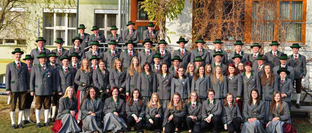 Eine Gruppe von Menschen, wahrscheinlich ein Chor, in traditionellen Outfits mit grünen Hüten, posiert für ein Foto vor einem Gebäude.