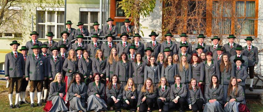 Eine Gruppe von Menschen, wahrscheinlich ein Chor, in traditionellen Outfits mit grünen Hüten, posiert für ein Foto vor einem Gebäude.