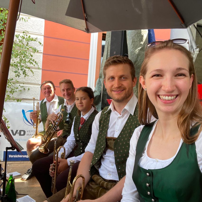 Eine Gruppe von Musikern, gekleidet in traditionelle grüne Westen und weiße Hemden, lächelt und posiert für ein Foto unter einem Regenschirm. Hinter ihnen ist ein Gebäude mit einer orangefarbenen Wand und einem weißen Banner zu sehen.
