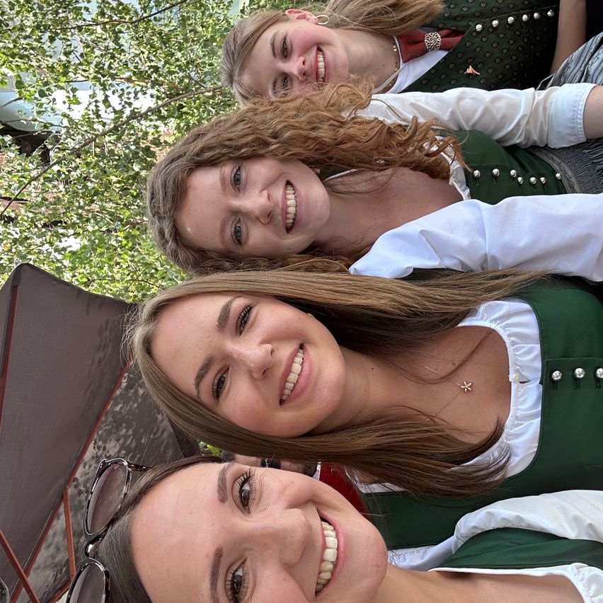 Vier Frauen in traditioneller deutscher Kleidung, lächelnd und posierend für ein Foto in einer Außenumgebung mit Bäumen und einem Baldachin im Hintergrund.