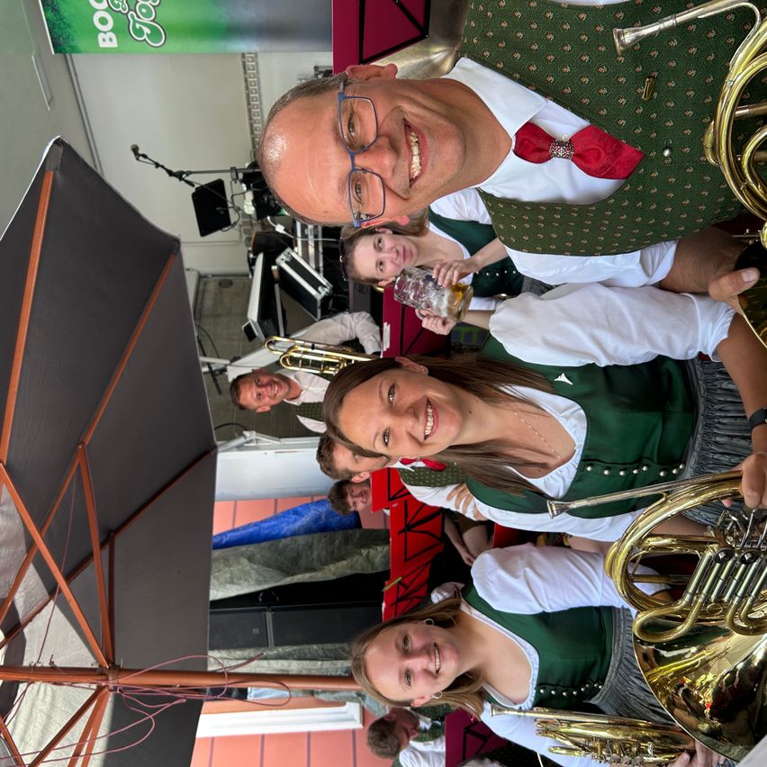 Eine Gruppe von Musikern in traditioneller Kleidung, die lächelnd für ein Foto posieren. Der Mann vorne trägt eine grüne Weste mit einer roten Fliege. Hinter ihnen hält eine Frau ein Glas Bier. Im Hintergrund sind Musikinstrumente zu sehen.
