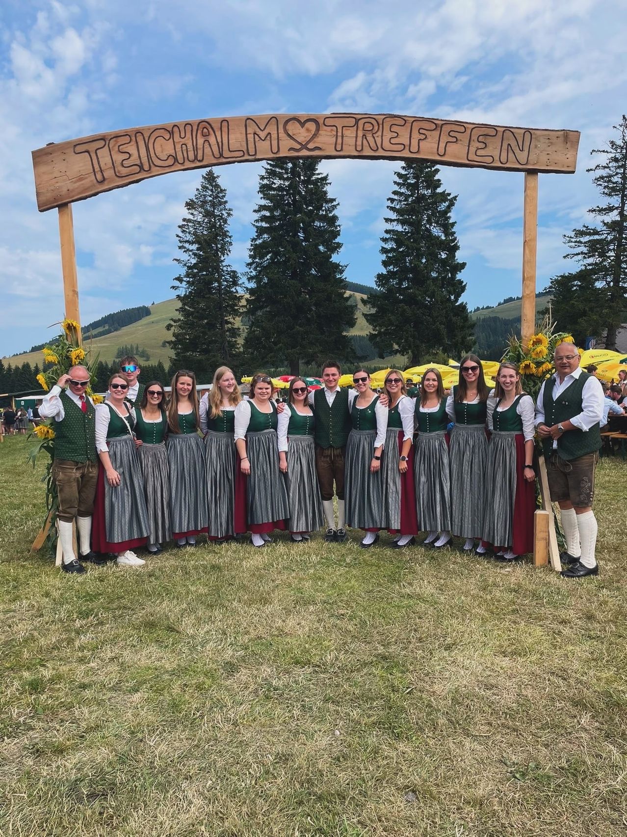 Eine Gruppe von Menschen in traditioneller deutscher Kleidung posiert für ein Foto vor einem Holzbogen mit den Worten Teichalm Treffen.
