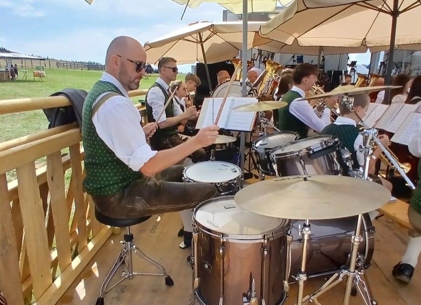 Ein Mann in einem grünen Vest spielt auf einer Bühne Schlagzeug mit anderen Musikern unter Schirmen, mit einem Grasfeld im Hintergrund.