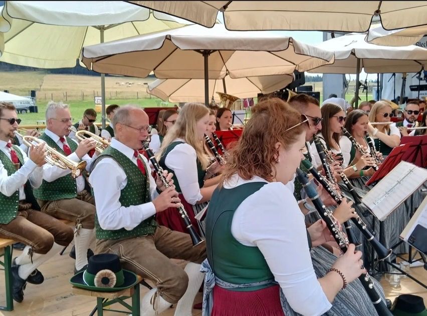 Eine Gruppe von Musikern in traditioneller Kleidung spielt verschiedene Blasinstrumente, darunter Trompeten und Klarinetten, unter großen Schirmen in einer Außenumgebung.