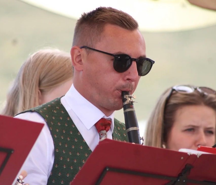 Ein Mann in einem grünen Weste und roter Krawatte spielt Klarinette vor einem roten Notenständer. Hinter ihm beobachtet eine Frau mit Sonnenbrille auf dem Kopf.