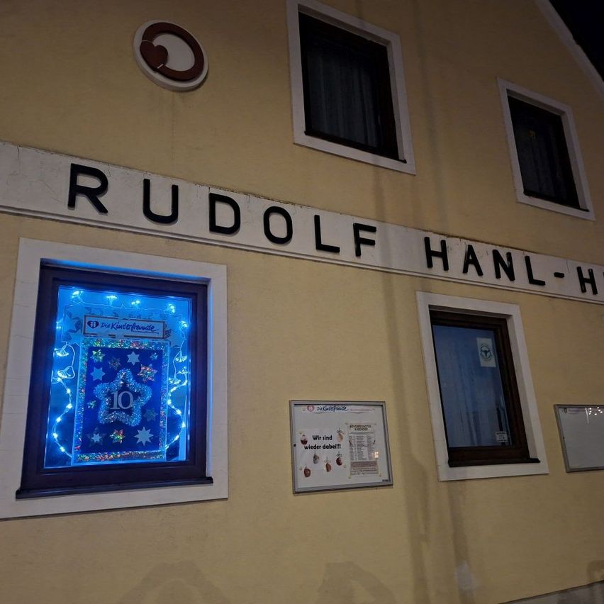 A building with the name Rudolf Hanl-Haus. A window with blue lights shows a star with the number 10. A sign with a red circle and logo is next to it.