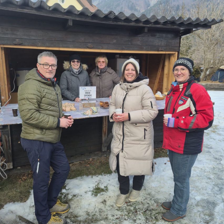 Fünf Erwachsene stehen vor einem verschneiten Chalet und lächeln für ein Foto. Sie halten Tassen und einige haben Essen. Ein Schild sagt 'Freie Spenden'.