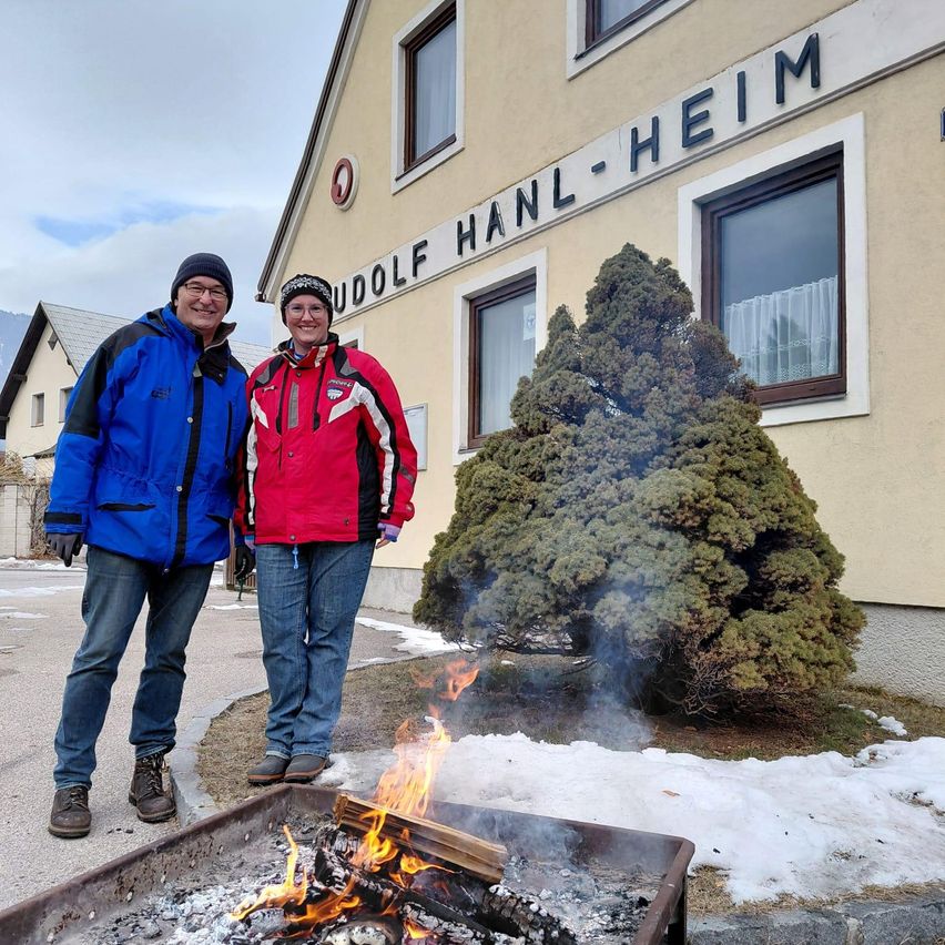 Zwei Personen in Wintermänteln stehen vor einem Gebäude mit dem Schild 'UDOLF HANL-HEIM'. Sie sind in der Nähe eines Feuerkorbs mit brennendem Holz. Die Umgebung ist verschneit.