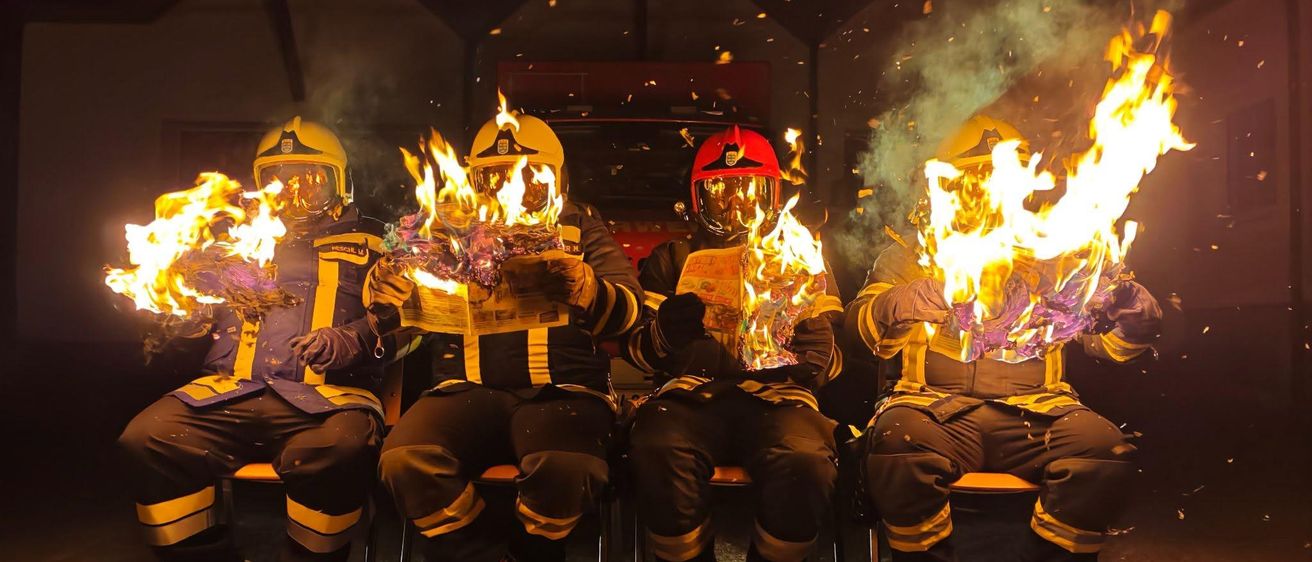 Fünf Feuerwehrleute in voller Ausrüstung sitzen auf Stühlen, mit brennenden Flammen auf ihren Knien, möglicherweise während einer Trainingsübung.